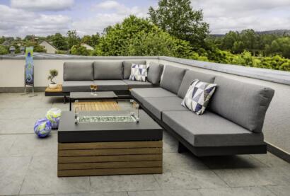 Moderne Außenterrasse in der Ferienwohnung Die Blaue Pause 42 mit einem grauen Sektionssofa, geometrischen Kissen, einem Tisch mit Feuerstelle, zwei Weingläsern und bunten Dekokugeln, alles umgeben von Grünpflanzen unter einem bewölkten Himmel.