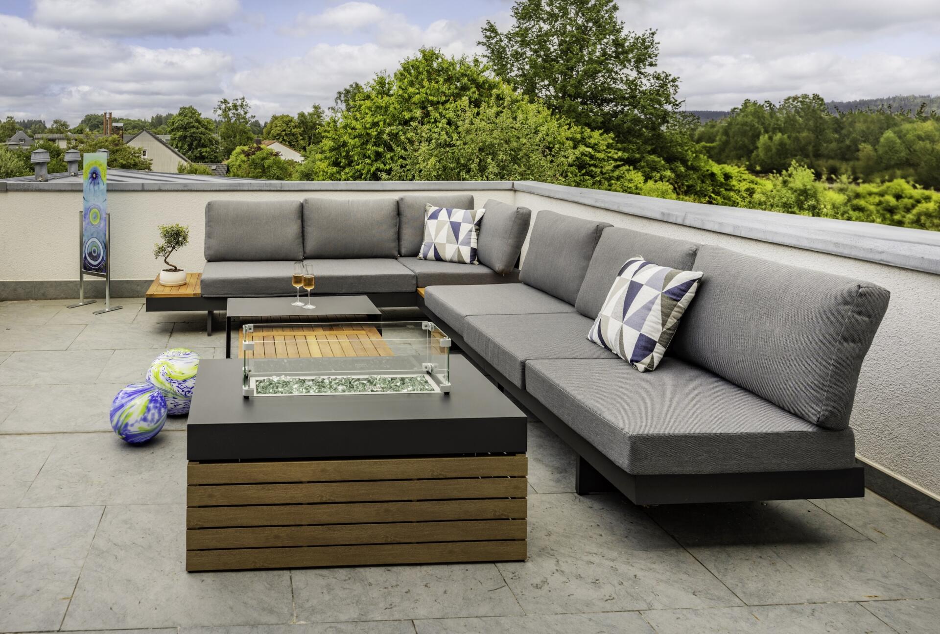 Moderne Außenterrasse in der Ferienwohnung Die Blaue Pause 42 mit einem grauen Sektionssofa, geometrischen Kissen, einem Tisch mit Feuerstelle, zwei Weingläsern und bunten Dekokugeln, alles umgeben von Grünpflanzen unter einem bewölkten Himmel.