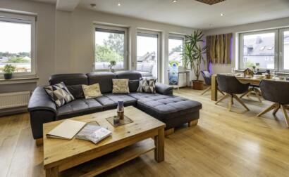 Modernes Wohnzimmer in der Ferienwohnung Die Blaue Pause 42 mit einem grauen Sektionssofa, einem Couchtisch aus Holz, einem Essbereich für vier Personen, großen Fenstern, einer Topfpflanze und einem hellen Holzfußboden. Natürliches Licht erhellt den Raum.