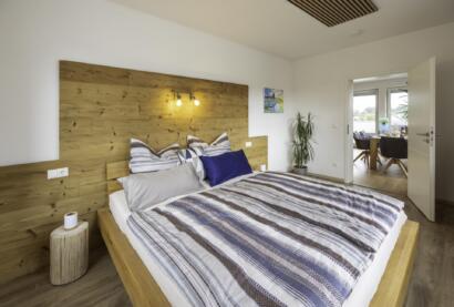 Modernes Schlafzimmer in der Ferienwohnung Die Blaue Pause 42 mit einem großen Bett mit gestreiftem Bettzeug, einem Kopfteil aus Holz und kleinen Nachttischen. Der helle Raum öffnet sich zu einem angrenzenden Raum mit Pflanzen und einem schönen Blick nach draußen.